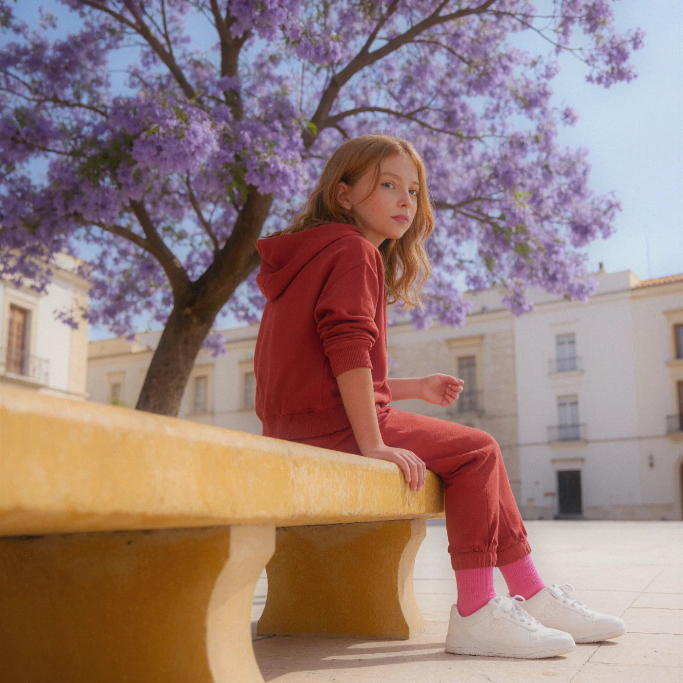 A child sitting on a stone bench in a Mediterranean plaza, mid-sentence, talking to someone outside the frame.