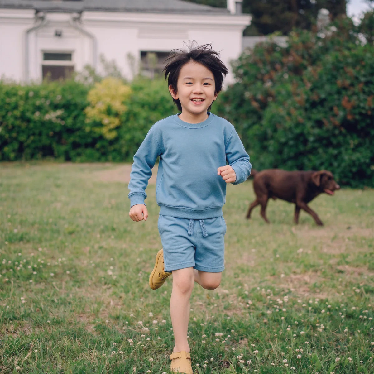 A kid running in a backyard wearing a sweatshirt and a short in blue shadow color.